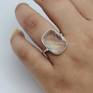 Op de afbeelding: Een zilveren ring met een heldere, gefacetteerde, rechthoekige edelsteen. De ring wordt aan een vinger gedragen.