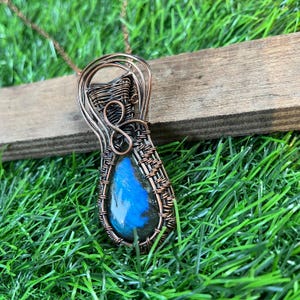 以下が含まれることがあります： 青いラブラドライトの宝石を特徴とする、銅線で包まれたペンダント。ペンダントは涙滴型で、編まれたワイヤーのデザインが施されています。