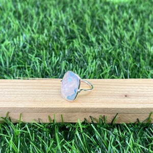 Puede incluir: Un anillo de plata con una piedra preciosa cuadrada e iridiscente. El anillo est&aacute; colocado sobre una superficie de madera.