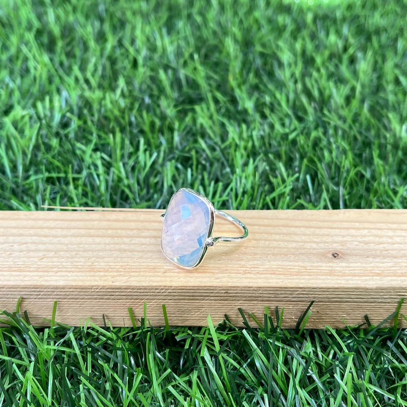 Puede incluir: Un anillo de plata con una piedra preciosa cuadrada e iridiscente. El anillo est&aacute; colocado sobre una superficie de madera.