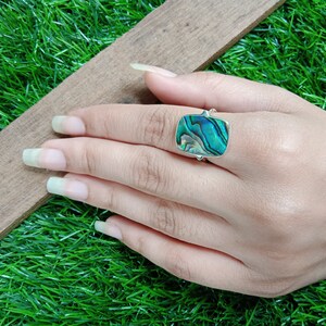 May include: A silver ring with a rectangular abalone shell inlay. The shell has a green, blue, and iridescent pattern.