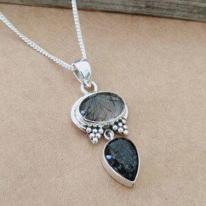May include: A silver necklace with a pendant featuring two black stones. The larger stone is oval-shaped and has a clear, cloudy appearance with black inclusions. The smaller stone is teardrop-shaped and has a more solid black color.