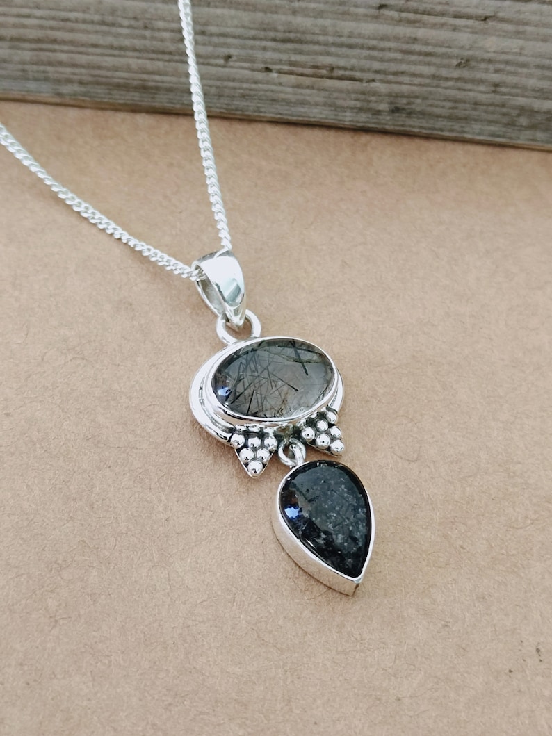 May include: A silver necklace with a pendant featuring two black stones. The larger stone is oval-shaped and has a clear, cloudy appearance with black inclusions. The smaller stone is teardrop-shaped and has a more solid black color.