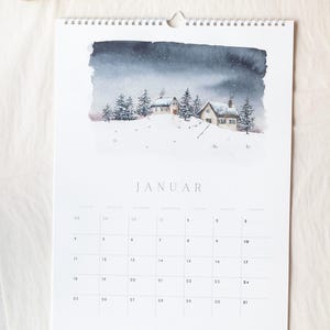 Könnte beinhalten: Ein Wandkalender für den Monat Januar mit einer Aquarellillustration von zwei Häusern auf einem verschneiten Hügel mit Tannenbäumen unter einem dunklen, bewölkten Himmel. Das Kalenderraster zeigt die Tage mit dem Text JANUAR am oberen Rand.