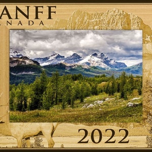 May include: A wooden picture frame with a carved image of mountains and a deer. The frame has a brown background with the words "BANFF CANADA" and "2022" printed on it.