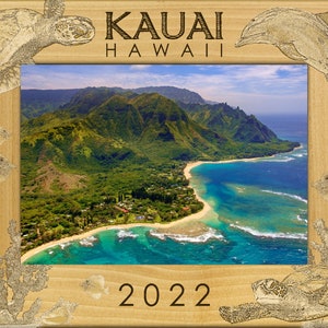 May include: A wooden picture frame with a carved design of sea turtles, dolphins, and fish. The frame has a photo of a beach scene with mountains in the background. The text "KAUAI HAWAII 2022" is carved into the frame.