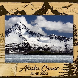May include: Wooden frame with an image of snow-capped mountains and a body of water under a blue sky. The frame has etched designs of a cruise ship, trees, and an eagle. Text at the bottom reads "Alaska Cruise June 2023."