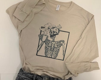 May include: A beige long-sleeved t-shirt with a black and white graphic of a skeleton drinking coffee. The shirt is paired with black denim shorts.