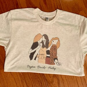 May include: A white t-shirt with a brown line drawing of three women hugging. The text "Peyton Brooke Haley" is printed below the drawing.