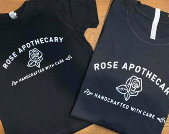 Puede incluir: Dos camisetas negras con texto blanco y un gráfico de una rosa. El texto dice "Rose Apothecary Handcrafted with Care".