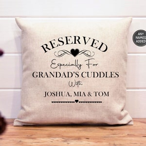 May include: A white pillow with black text that reads "RESERVED Especially For GRANDAD'S CUDDLES With JOSHUA, MIA & TOM". The text is surrounded by a decorative heart and scroll design.