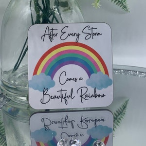 May include: A white square coaster with a rainbow and clouds. The text "After Every Storm Comes a Beautiful Rainbow" is printed on the coaster.