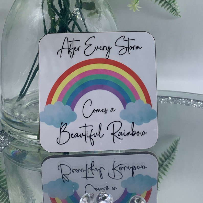 May include: A white square coaster with a rainbow and clouds. The text "After Every Storm Comes a Beautiful Rainbow" is printed on the coaster.