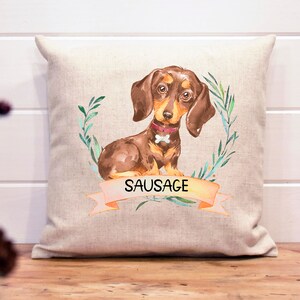 May include: A beige linen pillow with a watercolor illustration of a brown dachshund with a pink collar and a bone. The dog is surrounded by green leaves and the text "SAUSAGE" is on a peach-colored banner.
