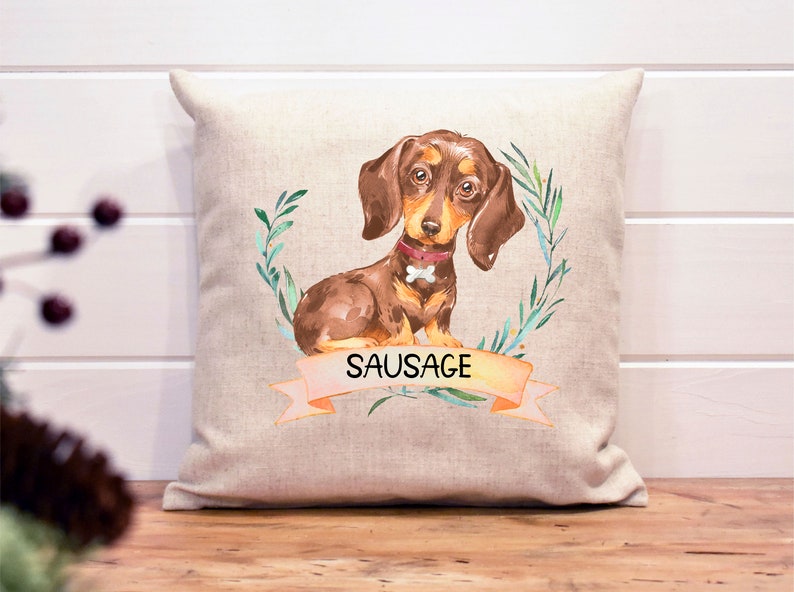 May include: A beige linen pillow with a watercolor illustration of a brown dachshund with a pink collar and a bone. The dog is surrounded by green leaves and the text "SAUSAGE" is on a peach-colored banner.