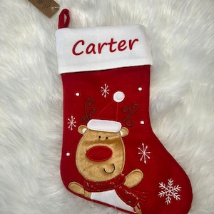 Pode incluir: Uma meia de Natal vermelha com um punho branco onde o nome Carter est&aacute; bordado em vermelho. A meia apresenta uma rena de pel&uacute;cia bege com um gorro de Pai Natal branco e um cachecol vermelho, rodeada por flocos de neve e pontos brancos.