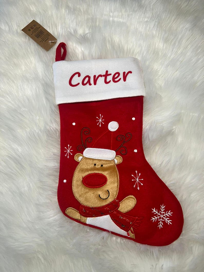 Pode incluir: Uma meia de Natal vermelha com um punho branco onde o nome Carter est&aacute; bordado em vermelho. A meia apresenta uma rena de pel&uacute;cia bege com um gorro de Pai Natal branco e um cachecol vermelho, rodeada por flocos de neve e pontos brancos.