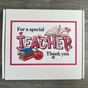 May include: White gift box with a pink and red border featuring the text "For a special TEACHER Thank you." The word TEACHER is in a colorful plaid pattern with floral accents. An open book, stack of books, and a red apple are also shown.