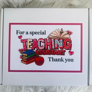 May include: A white box with a pink border featuring the text "For a special TEACHING ASSISTANT Thank you." The word "TEACHING" is in a colorful, patterned font. The image includes illustrations of books, flowers, and an apple.