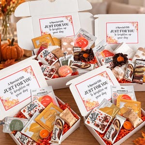 May include: Four white gift boxes filled with assorted items including Vitamin C face masks, Galaxy chocolate bars, small candles, and mugs with Hello Autumn text. Each box lid features a label reading A Special Gift JUST FOR YOU To brighten up your day.