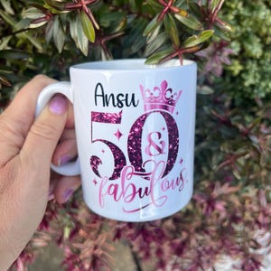 May include: White ceramic mug with the name "Ansu" and the text "50 & fabulous" in pink glitter lettering. A pink crown is above the number 50. The mug has a white handle and is surrounded by green and purple foliage.