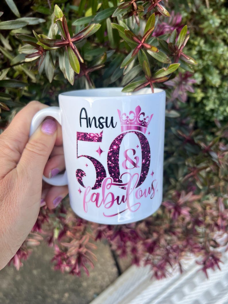 May include: White ceramic mug with the name "Ansu" and the text "50 & fabulous" in pink glitter lettering. A pink crown is above the number 50. The mug has a white handle and is surrounded by green and purple foliage.