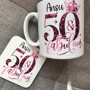 May include: White ceramic mug and coaster set with a pink and glittery design. The mug and coaster feature the name "Ansu", the number "50", the word "&", and the word "fabulous" in a script font. A pink crown is also present.
