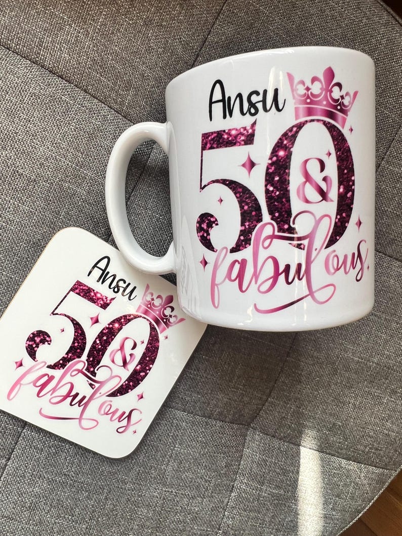 May include: White ceramic mug and coaster set with a pink and glittery design. The mug and coaster feature the name "Ansu", the number "50", the word "&", and the word "fabulous" in a script font. A pink crown is also present.