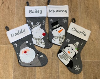 May include: Four gray Christmas stockings with white cuffs and hanging loops. Each stocking has a different design: Santa, a reindeer, a snowman, and a penguin. The names "Bailey", "Mummy", "Daddy", and "Charlie" are embroidered on the cuffs.