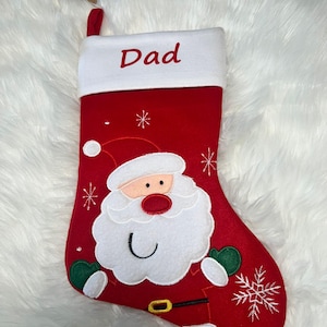 Pode incluir: Uma meia de Natal vermelha com um punho branco com a palavra Dad bordada em vermelho. A meia apresenta uma grande aplica&ccedil;&atilde;o do rosto do Pai Natal com barba branca, nariz vermelho e luvas verdes, acentuada por flocos de neve bordados.