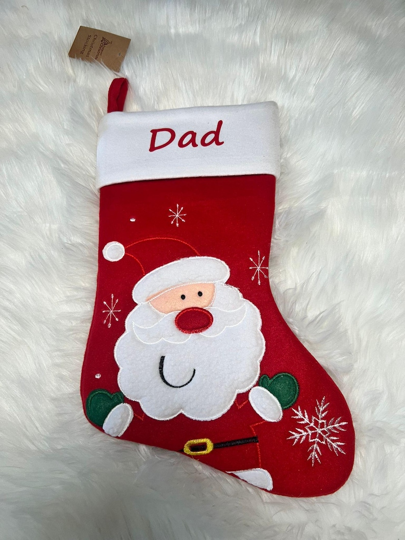 Pode incluir: Uma meia de Natal vermelha com um punho branco com a palavra Dad bordada em vermelho. A meia apresenta uma grande aplica&ccedil;&atilde;o do rosto do Pai Natal com barba branca, nariz vermelho e luvas verdes, acentuada por flocos de neve bordados.