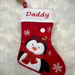Pode incluir: Uma meia de Natal vermelha com um punho branco bordado com a palavra Daddy em vermelho. A meia apresenta uma aplica&ccedil;&atilde;o de um pinguim com gorro e cachecol vermelhos, acentuada com flocos de neve bordados a branco e um pequeno ramo de azevinho.