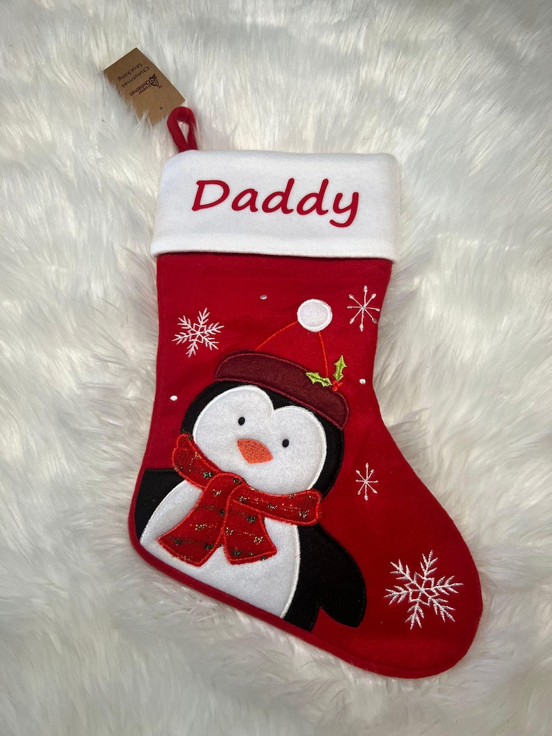 Pode incluir: Uma meia de Natal vermelha com um punho branco bordado com a palavra Daddy em vermelho. A meia apresenta uma aplica&ccedil;&atilde;o de um pinguim com gorro e cachecol vermelhos, acentuada com flocos de neve bordados a branco e um pequeno ramo de azevinho.