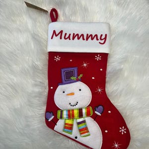 Pode incluir: Uma meia de Natal vermelha com um punho branco onde a palavra Mummy est&aacute; bordada a vermelho. A meia apresenta um boneco de neve aplicado com uma cartola roxa e um cachecol &agrave;s riscas multicolorido, rodeado por flocos de neve brancos bordados.