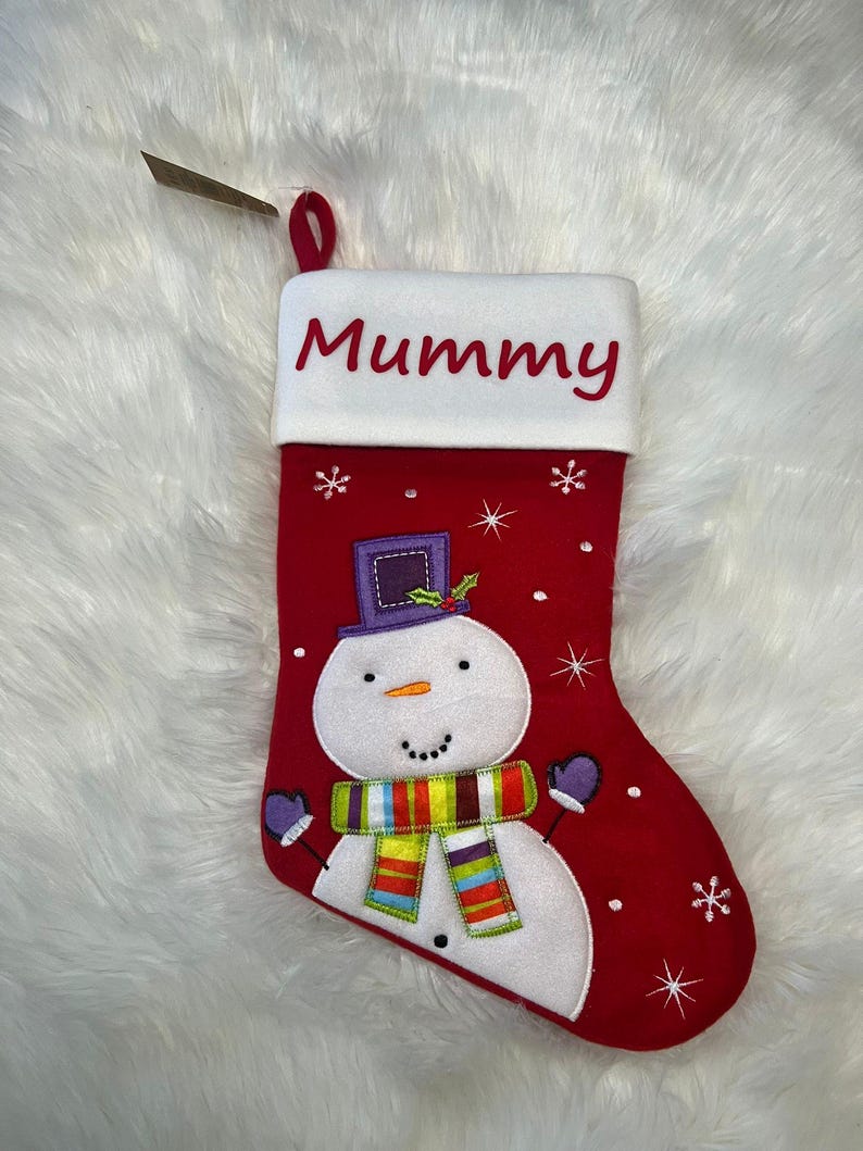 Pode incluir: Uma meia de Natal vermelha com um punho branco onde a palavra Mummy est&aacute; bordada a vermelho. A meia apresenta um boneco de neve aplicado com uma cartola roxa e um cachecol &agrave;s riscas multicolorido, rodeado por flocos de neve brancos bordados.