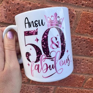 May include: White ceramic mug with the name "Ansu", the number "50", the word "fabulous", and a crown design in pink glitter. The mug has a handle and is designed for beverages.