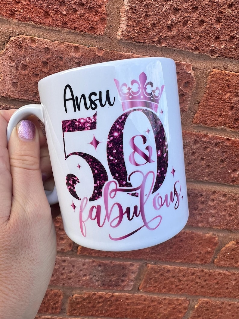 May include: White ceramic mug with the name "Ansu", the number "50", the word "fabulous", and a crown design in pink glitter. The mug has a handle and is designed for beverages.