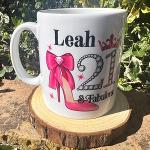 May include: White ceramic mug with the name "Leah", the number "21", and the words "& Fabulous" printed on it. The mug features a pink high heel, a pink bow, and a crown design. The mug is sitting on a wooden coaster.