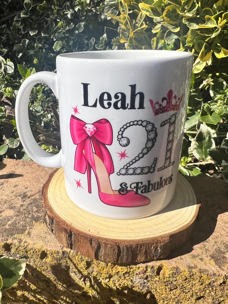 May include: White ceramic mug with the name "Leah", the number "21", and the words "& Fabulous" printed on it. The mug features a pink high heel, a pink bow, and a crown design. The mug is sitting on a wooden coaster.