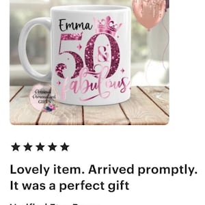 May include: White ceramic mug with the text "Emma 50 & fabulous" in pink glitter lettering. The mug has a handle and a crown design. Two balloons are in the background.