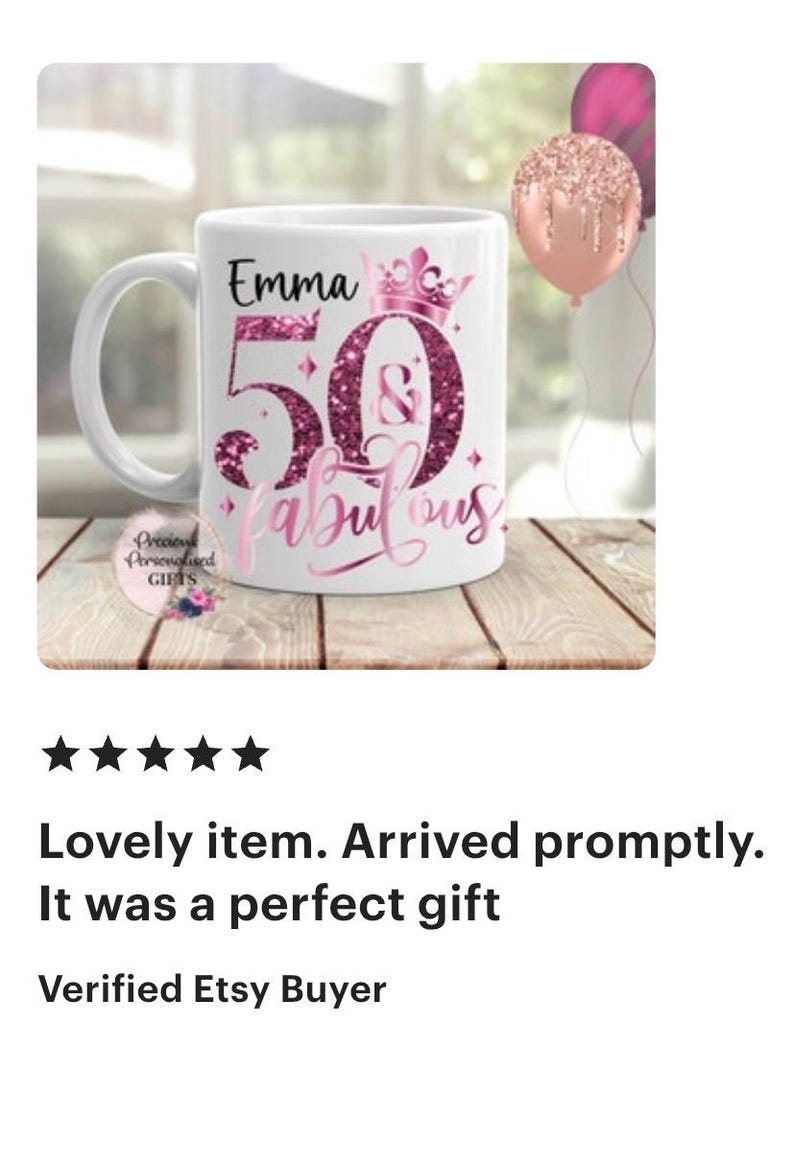 May include: White ceramic mug with the text "Emma 50 & fabulous" in pink glitter lettering. The mug has a handle and a crown design. Two balloons are in the background.
