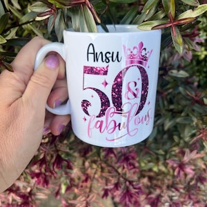May include: White ceramic mug with a handle, featuring the name "Ansu" in black script above the number "50" and the word "fabulous" in pink script. The number "50" is filled with pink glitter, and a pink crown is above the number.