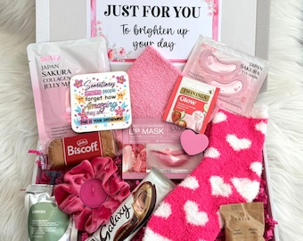 Pode incluir: Uma caixa de presente contendo uma máscara de gelatina de colágeno Japan Sakura, um cartão quadrado com texto inspirador, uma meia rosa felpuda com corações brancos, um saquinho de chá Twinings Glow, um biscoito Biscoff, uma barra de chocolate Galaxy e um elástico.