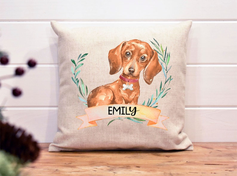 May include: A beige linen pillow with a watercolor illustration of a dachshund dog with a pink collar and the name "Emily" on a banner.