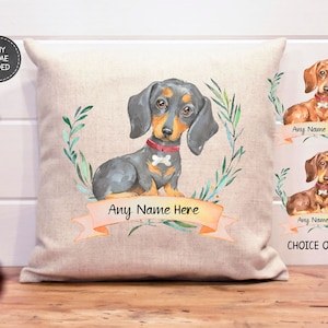 May include: A personalized pillow featuring a watercolor illustration of a dachshund dog with a red collar and a bone charm. The pillow is beige with a green leafy wreath around the dog and a pink ribbon with the text "Any Name Here".