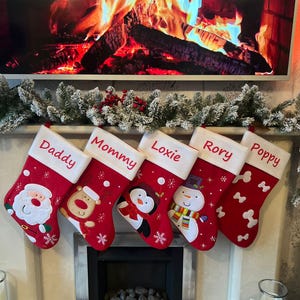 Pode incluir: Cinco meias de Natal vermelhas com punhos brancos penduradas em fila. As meias apresentam nomes bordados: Daddy, Mommy, Loxie, Rory e Poppy. Os designs incluem o Pai Natal, uma rena, um pinguim, um boneco de neve e formas de ossos brancos.