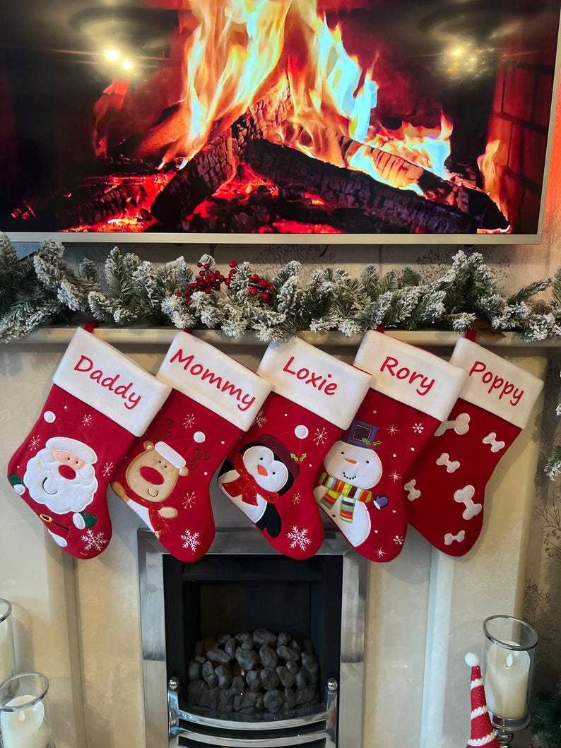 Pode incluir: Cinco meias de Natal vermelhas com punhos brancos penduradas em fila. As meias apresentam nomes bordados: Daddy, Mommy, Loxie, Rory e Poppy. Os designs incluem o Pai Natal, uma rena, um pinguim, um boneco de neve e formas de ossos brancos.