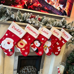May include: Six red Christmas stockings with white cuffs are displayed above a fireplace. Each stocking is personalised with a name: Daddy, Mommy, Loxie, Rory, and Poppy. The stockings are decorated with festive characters like Santa and a snowman.