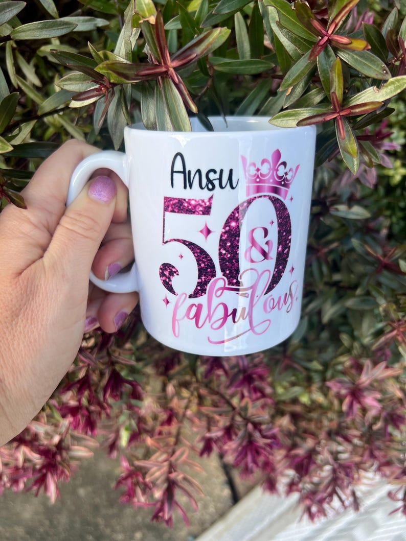 May include: White ceramic mug with a handle, featuring the name "Ansu" in black script above the number "50" and the word "fabulous" in pink script. The number "50" is filled with pink glitter, and a pink crown is above the number.