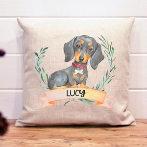 May include: A beige linen pillow with a watercolor illustration of a black and tan dachshund with a red collar and a bone charm. The dog is surrounded by green leaves and the name "LUCY" is written on a pink banner.
